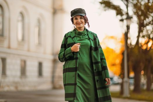 Outdoor fashion portrait of an elegant fashionable brunette woman, model in a stylish cap, green dress, posing at sunset in a European city in autumn