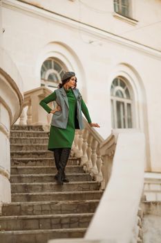 Outdoor fashion portrait of an elegant fashionable brunette model in a beret, green dress and a gray waistcoat posing at sunset in a european city in autumn