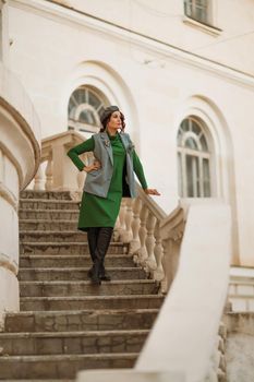 Outdoor fashion portrait of an elegant fashionable brunette model in a beret, green dress and a gray waistcoat posing at sunset in a european city in autumn