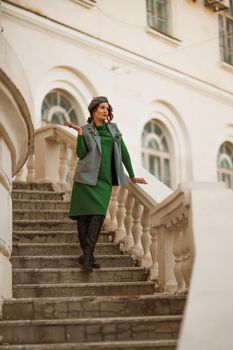 Outdoor fashion portrait of an elegant fashionable brunette model in a beret, green dress and a gray waistcoat posing at sunset in a european city in autumn