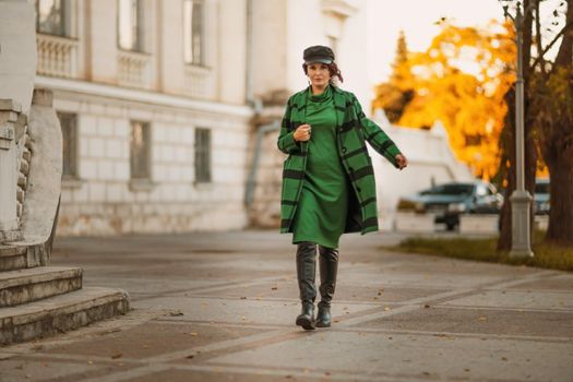 Outdoor fashion portrait of an elegant fashionable brunette woman, model in a stylish cap, green dress, posing at sunset in a European city in autumn