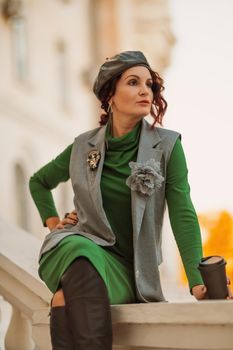 Outdoor fashion portrait of an elegant fashionable brunette model in a beret, green dress and a gray waistcoat posing at sunset in a european city in autumn