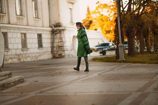 Outdoor fashion portrait of an elegant fashionable brunette woman, model in a stylish cap, green dress, posing at sunset in a European city in autumn