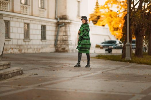 Outdoor fashion portrait of an elegant fashionable brunette woman, model in a stylish cap, green dress, posing at sunset in a European city in autumn