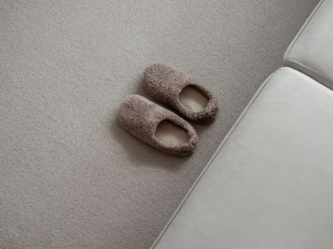 Soft fur warm slippers on carpet near sofa in simple scandinavian home. Pair of trendy fashion brown warm slippers on floor, copy space, top view