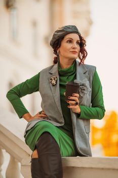 Outdoor fashion portrait of an elegant fashionable brunette model in a beret, green dress and a gray waistcoat posing at sunset in a european city in autumn