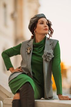 Outdoor fashion portrait of an elegant fashionable brunette model in a beret, green dress and a gray waistcoat posing at sunset in a european city in autumn