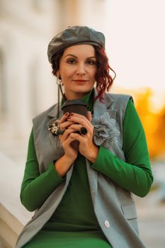 Outdoor fashion portrait of an elegant fashionable brunette model in a beret, green dress and a gray waistcoat posing at sunset in a european city in autumn