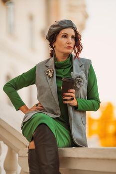 Outdoor fashion portrait of an elegant fashionable brunette model in a beret, green dress and a gray waistcoat posing at sunset in a european city in autumn