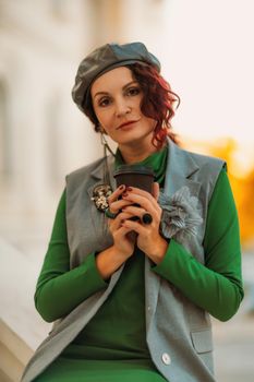 Outdoor fashion portrait of an elegant fashionable brunette model in a beret, green dress and a gray waistcoat posing at sunset in a european city in autumn