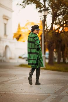 Outdoor fashion portrait of an elegant fashionable brunette woman, model in a stylish cap, green dress, posing at sunset in a European city in autumn