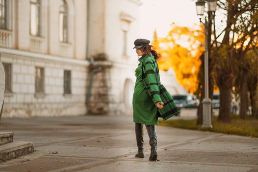 Outdoor fashion portrait of an elegant fashionable brunette woman, model in a stylish cap, green dress, posing at sunset in a European city in autumn