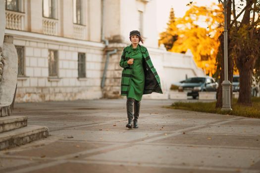 Outdoor fashion portrait of an elegant fashionable brunette woman, model in a stylish cap, green dress, posing at sunset in a European city in autumn