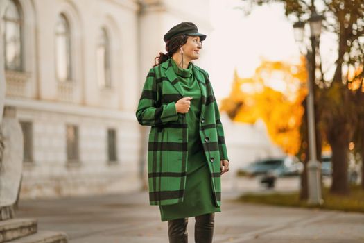 Outdoor fashion portrait of an elegant fashionable brunette woman, model in a stylish cap, green dress, posing at sunset in a European city in autumn