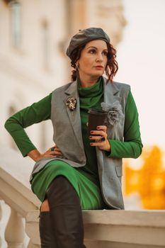 Outdoor fashion portrait of an elegant fashionable brunette model in a beret, green dress and a gray waistcoat posing at sunset in a european city in autumn