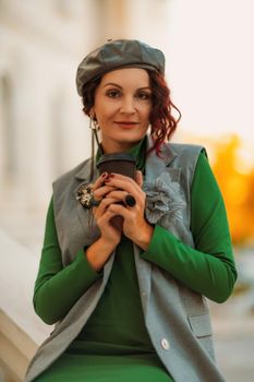 Outdoor fashion portrait of an elegant fashionable brunette model in a beret, green dress and a gray waistcoat posing at sunset in a european city in autumn