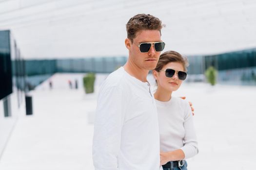 Smiling beautiful woman and her handsome boyfriend. Woman in casual summer jeans. Happy cheerful couple in sunglasses walking business district. Couple posing on the street modern building background