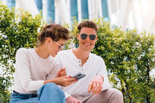 Young modern couple use smartphone sitting bench outdoor in office district. Beautiful woman and handsome man in sunglasses holding smartphone in hand looking device screen and smile.