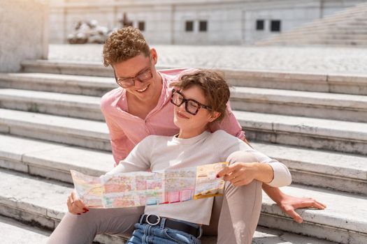 Tourist Couple With Map In Capital City Europe. Happy Young People Using Map, Traveling On Summer Vacations. Handsome Man And Beautiful Woman Having Trip Outdoors. High Quality Image. Sitting stairs