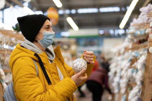 Christmas decoration shopping. Caucasian woman choose new year decor in shopping mall. Medium age female dressed medical mask and yellow winter parka buying Christmas ornament toys indoor store
