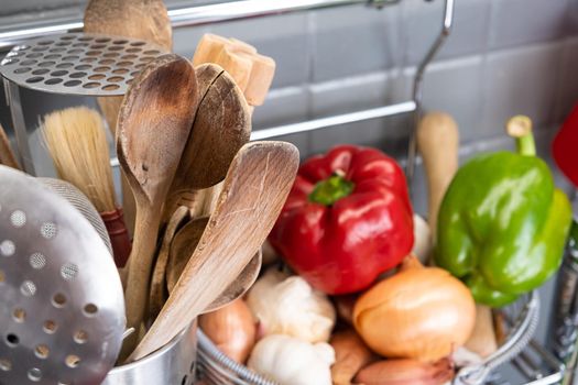 small basket in the kitchen next to the utensils with garlic, onions, shallots and red yellow and green peppers.