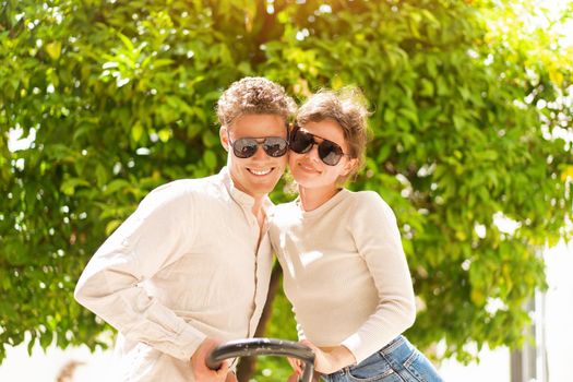 Smiling beautiful woman and her handsome boyfriend. Woman embrace summer day. Happy cheerful couple in sunglasses walking old city park. Couple posing on the street near green tree