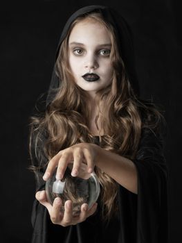 Little girl in Halloween witch costume and dark make-up holding crystal ball isolated on black background