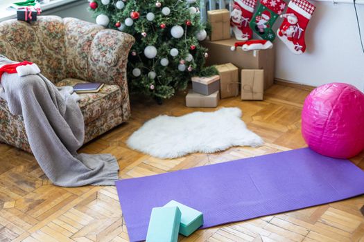Empty space in fitness center with big windows and natural wooden floor. Unrolled yoga mat on the floor, no people. Decorated christmas tree