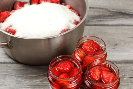 Bottles with homemade pickled strawberries, steel pot with more berries covered with sugar in background. Home made preserved fruit preparation.