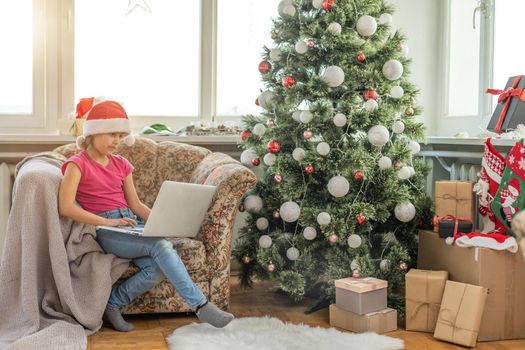 Christmas tree in a large living room. Little girl plays near the Christmas tree. A girl with a laptop communicates with friends.