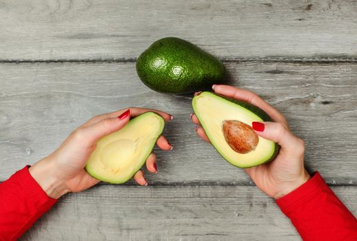 Tabletop view, woman hand holding two halves of avocado in each hand, one more whole pear placed on gray wooden desk above.