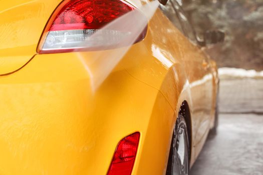 Yellow car rear light and bumper washed in self serve carwash, water jet spraying on gloss surface.