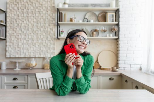 In love and happy young beautiful woman sends and receives love messages on the red phone from a loved one, from a boyfriend. Standing in the kitchen at home in glasses and a green shirt.