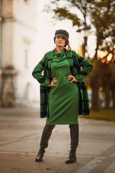 Outdoor fashion portrait of an elegant fashionable brunette woman, model in a stylish cap, green dress, posing at sunset in a European city in autumn