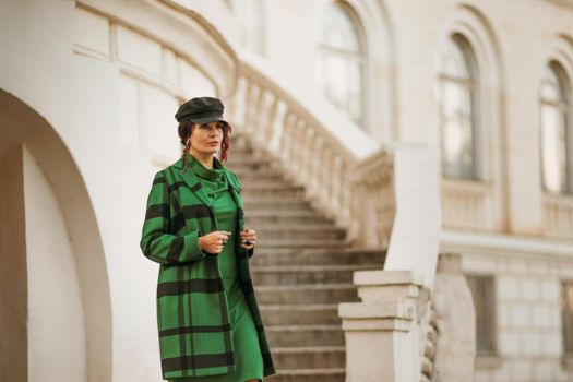 Outdoor fashion portrait of an elegant fashionable brunette woman, model in a stylish cap, green dress, posing at sunset in a European city in autumn