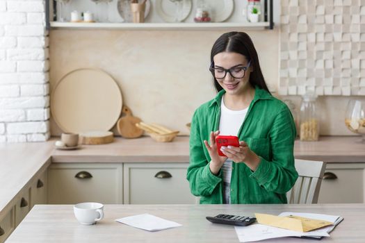 Satisfied young beautiful businesswoman working at home in kitchen with red phone, documents and calculator. Sitting at the table near the window in glasses and a green shirt.