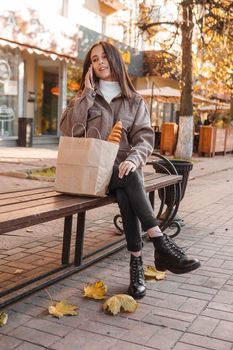 A stylish brunette woman walks around the autumn city. The brunette is sitting on a bench outside with a glass of coffee and a phone, holding a craft bag with a baguette.