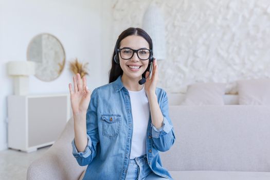 Webinar. Young beautiful business woman in glasses and headphones sits on the sofa at home and conducts a webinar, online training on camera, consults, tells, gestures with hands, smiles.