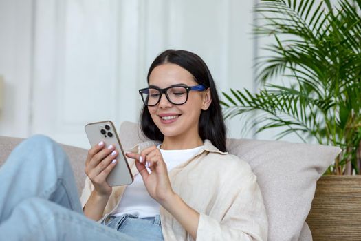 Happy young student girl in glasses sitting on sofa at home, holding phone, chatting and chatting with friends, smiling.