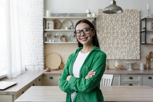 Portrait of a young beautiful brunette woman in glasses and a green shirt. Standing in the kitchen at home, looking at the camera, smiling, folded her arms over her chest.