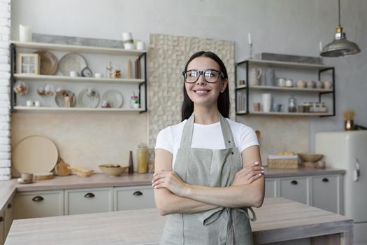 Portrait of a young beautiful woman cook, confectioner. She is standing in a beige apron and glasses in the kitchen, her arms crossed. He looks at the camera and smiles.