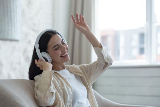 Beautiful young woman listening to music in white headphones. He sits at home on the sofa with his eyes closed, dances, sings, waves his hands.