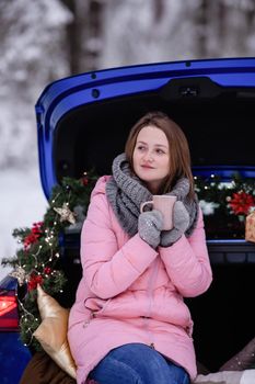 A woman in winter clothes drinks a hot drink sitting in a car decorated in a New Year's way. Traveling by car through the forest. A trip before Christmas.