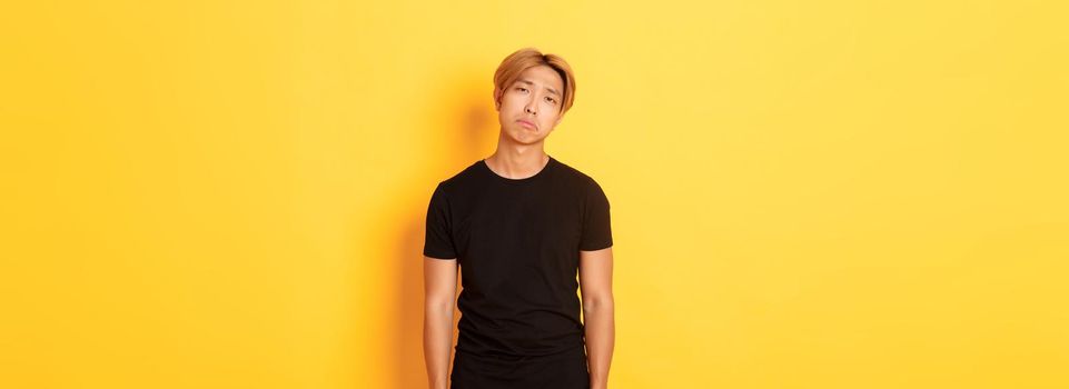 Portrait of exhausted and sad asian guy looking sad, standing drained over yellow background.