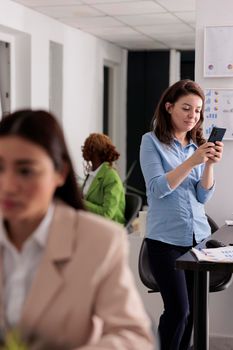 Smiling employee sending text messages on smartphone, standing near workplace desk, chatting on social media in coworking space. Office worker chatting on mobile phone, typing on cellphone