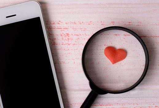 heart under a magnifying glass, lies near a white mobile phone. Concept of mobile dating sites, search for love. High quality photo