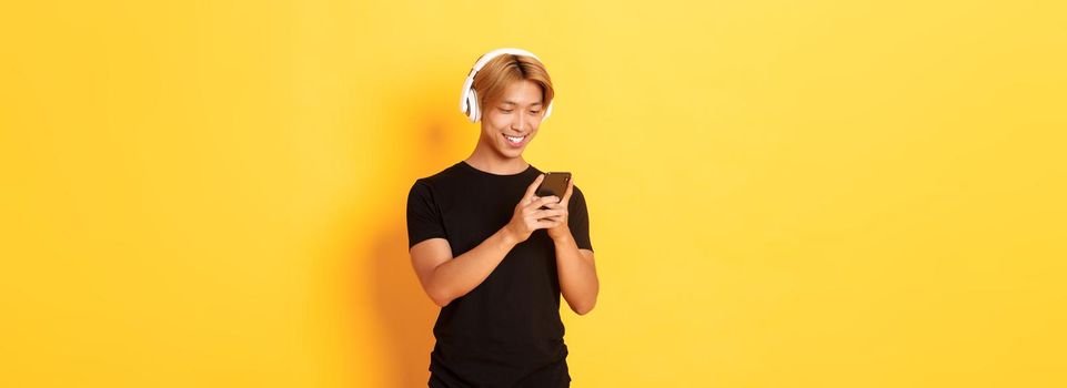 Young smiling asian guy using smartphone and listening music or podcast in wireless headphones, standing yellow background.