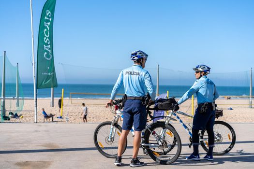 Portugal, Carcavelos April 2022 Two policeman patrolling seaside promenade on bicycles. People are sunburning on the city public beach on the Atlantic shore. Bike police patrol on the ocean beach