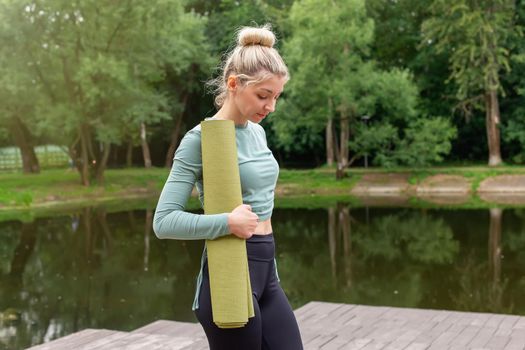A beautiful slender blonde, holding a green gym mat in her hands, stands in the park by the pond in summer, looking down. Copy space