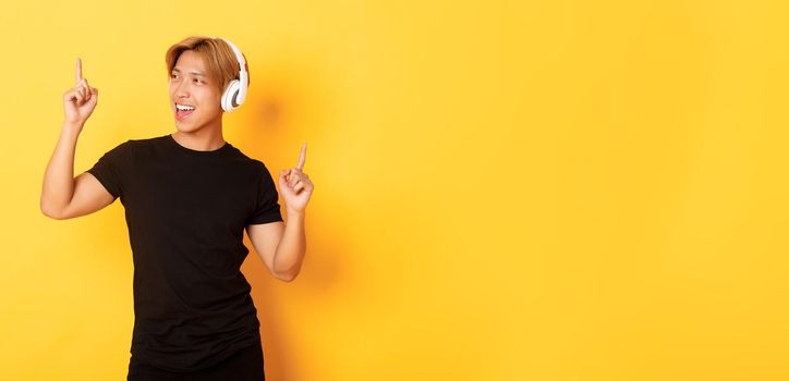 Joyful handsome asian guy with blond hair, singing along and dancing as listening to music in wireless headphones, standing yellow background.